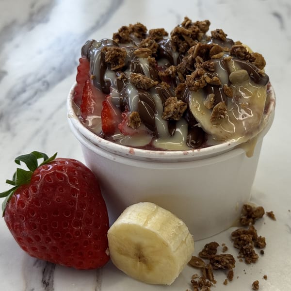Açaí bowl with strawberries, bananas, granola, chocolate, and condensed milk drizzles, with fruit on the side.