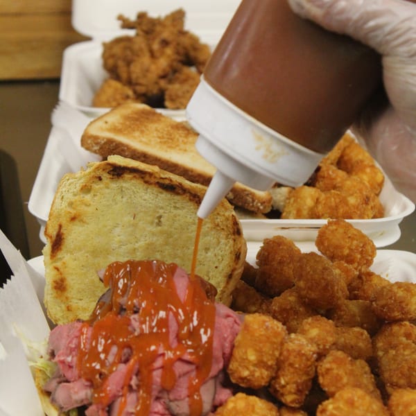 Gloved hand squeezing sauce onto a roast beef sandwich with tater tots, fried chicken, and toast in takeout containers.