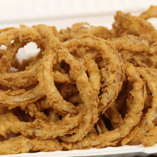 Golden-brown crispy onion rings piled in a white takeout container, with a hint of red sauce.