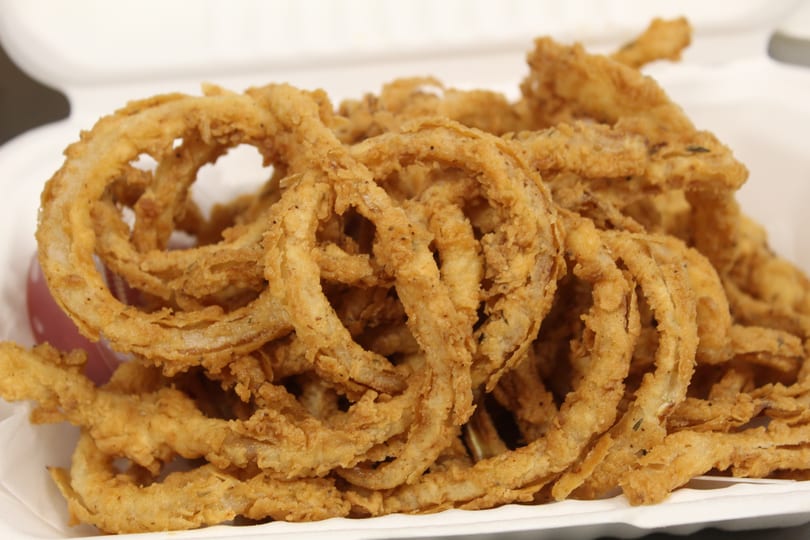 Golden-brown crispy onion rings piled in a white takeout container, with a hint of red sauce.