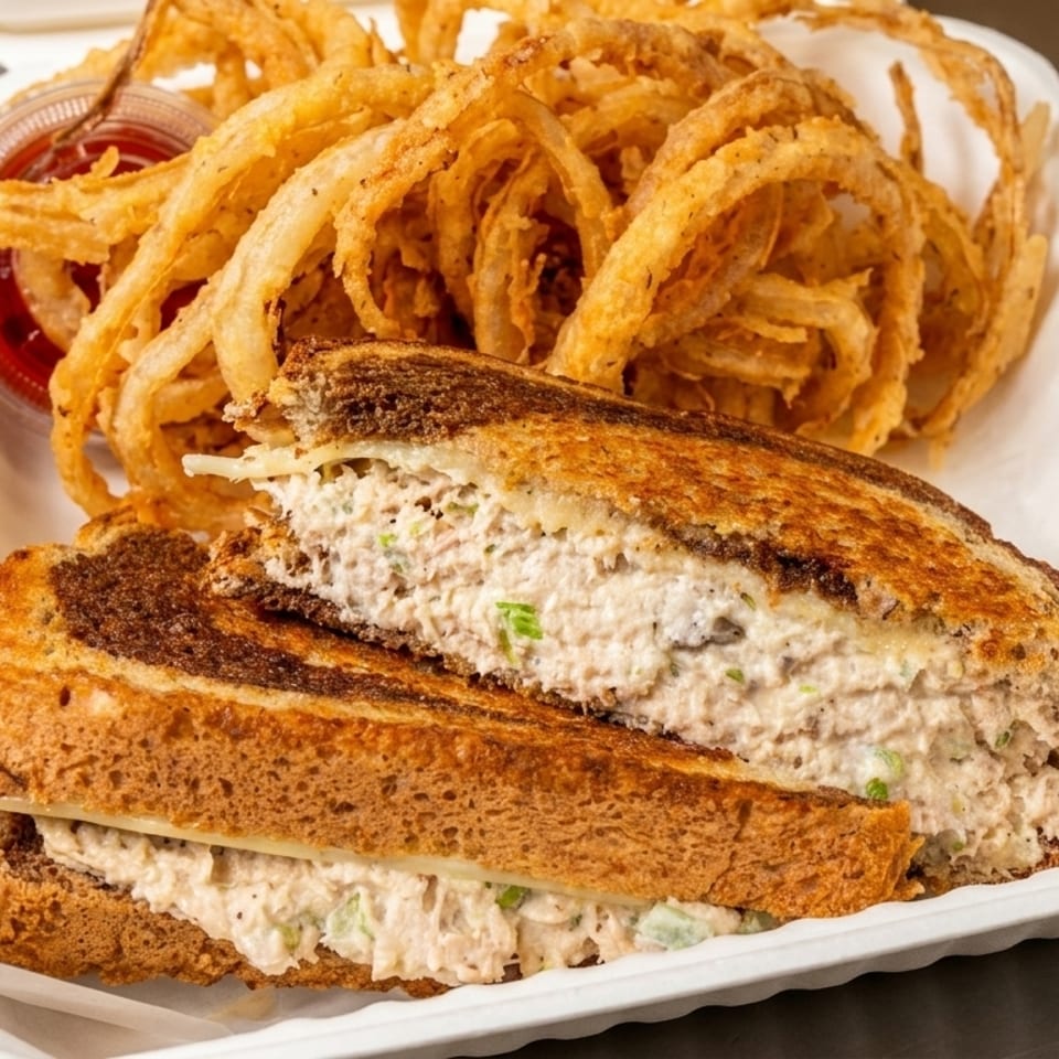 Tuna melt sandwich on grilled rye bread with a side of crispy onion rings and ketchup.